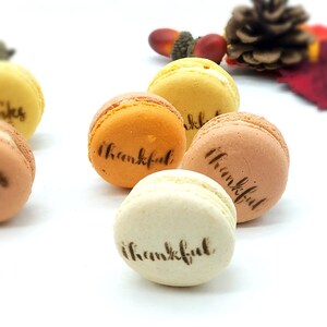 May include: A collection of colorful macarons in shades of yellow, orange, and brown, each with the word "thankful" or "happy thanks giving" printed on top. The macarons are arranged on a white surface, with fall-themed decorations in the background.