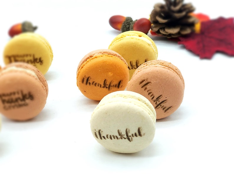May include: A collection of colorful macarons in shades of yellow, orange, and brown, each with the word "thankful" or "happy thanks giving" printed on top. The macarons are arranged on a white surface, with fall-themed decorations in the background.