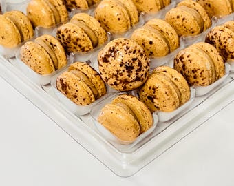 May include: A clear plastic container filled with a variety of beige macarons with brown chocolate sprinkles. The macarons are arranged in rows and columns.