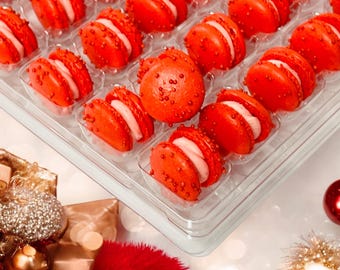 May include: A clear plastic container filled with red macarons with white frosting. The macarons are decorated with red sprinkles. The container is sitting on a white surface with gold and red decorations.