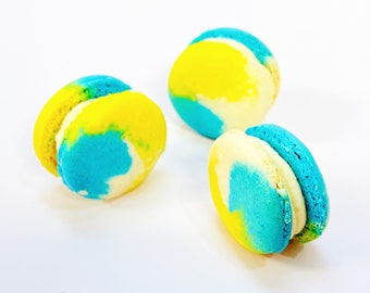 Può includere: Tre macarons blu e gialli con glassa bianca. I macarons sono disposti a forma di triangolo su una superficie bianca.