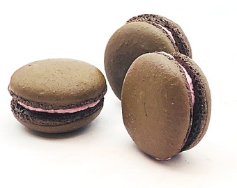 Può includere: Tre macarons al cioccolato con ripieno rosa. I macarons sono disposti in fila su una superficie bianca.