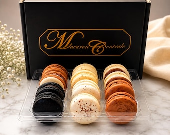 May include: A black rectangular box with the gold text Macaron Centrale, containing a clear plastic tray of assorted macarons. The macarons are arranged in three rows of colors: black, cream with brown speckles, and shades of brown and tan.