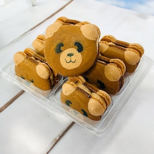 May include: Six brown macarons with a bear face design. The macarons are arranged in a clear plastic container.