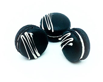 May include: Three dark macarons with white drizzle against a white backdrop. The macarons are round with a white filling and a white drizzle pattern on top. The cookies are a dark colour, possibly black or dark chocolate.