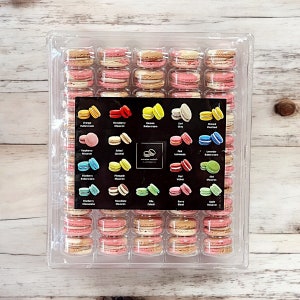 May include: A clear plastic container filled with 36 colorful macarons. The macarons are arranged in rows and columns, with each macaron having a different flavor. The flavors are listed on a black label in the center of the container. The flavors include: Orange Buttercream, Strawberry Macaron, Banana Buttercream, Earl Grey, Almond Overload, Raspberry Macaron, Salted Caramel, Pink Lemonade, Lavender Buttercream, Blueberry Buttercream, Pineapple Macaron, Red Velvet, Green Macaron,  Macaron Cheesecake, Mango Macaron, Cola, Berry Blend, Apple Macaron.