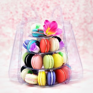 May include: A clear plastic tiered stand filled with colorful macarons. The stand has four tiers and is decorated with a pink flower.