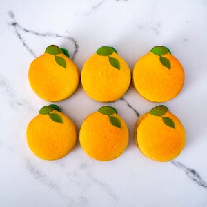 May include: Six orange-colored macarons, each with a green leaf detail, arranged on a white marble surface. The macarons are round and have a smooth texture, resembling miniature oranges. The overall presentation is clean and visually appealing.