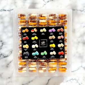 May include: A clear plastic container filled with colorful macarons. The macarons are arranged in rows and columns. A black card displays the macaron flavors, including Peach 'n Cream, Lychee Buttercream, and Grape Macaron. The macarons are a variety of colors.