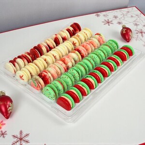 May include: A clear plastic tray filled with colorful macarons. Rows of red, white, pink, and green macarons are arranged in the tray. Two red Christmas ornaments are on the white surface, decorated with snowflakes.