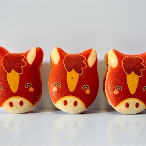 May include: Three red macarons, each decorated with a horse face design. The macarons have a red base with yellow and white accents, including a yellow nose and white ears. The macarons are arranged in a row against a white background.