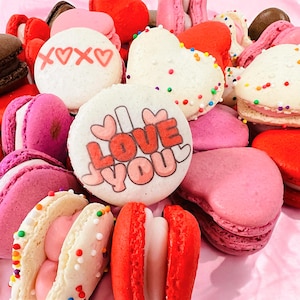 May include: A variety of heart-shaped and round macarons decorated with sprinkles, hearts, and the words "I Love You". The macarons are pink, white, and red.