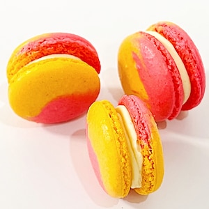 May include: Three yellow and pink macarons with white frosting. The macarons are arranged in a triangular shape on a white surface.
