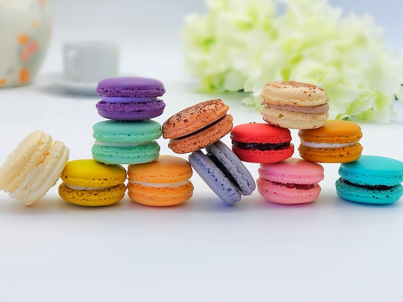 May include: A collection of colorful macarons, including purple, green, orange, yellow, brown, red, pink, and blue. The macarons are arranged in a semi-circle on a white surface.