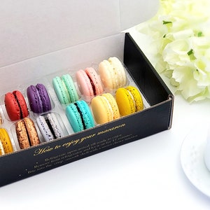 May include: A box of colorful macarons in a clear plastic tray. The box is black with gold lettering that says "How to enjoy your macaron". The macarons are arranged in rows of three. The macarons are in the colors of blue, green, pink, yellow, and orange.