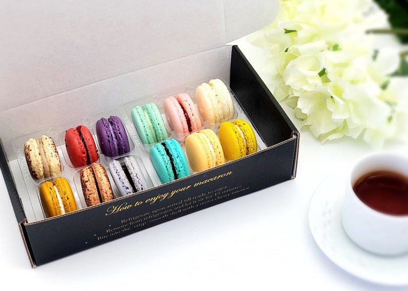 May include: A box of colorful macarons in a clear plastic tray. The box is black with gold lettering that says "How to enjoy your macaron". The macarons are arranged in rows of three. The macarons are in the colors of blue, green, pink, yellow, and orange.