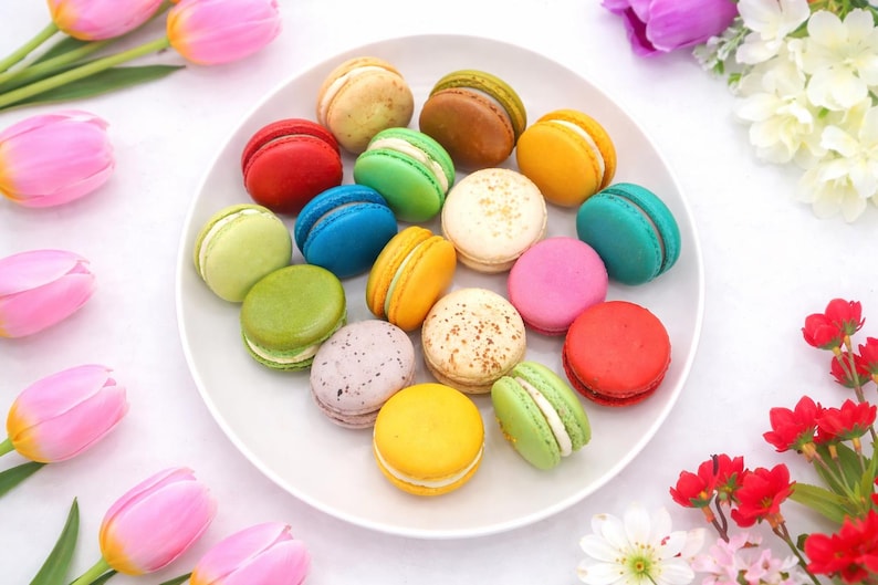 May include: A white plate filled with an assortment of colorful macarons in shades of red, blue, green, yellow, pink, brown, and white, some featuring speckled textures, arranged in a circular pattern surrounded by pink and red tulip flowers.