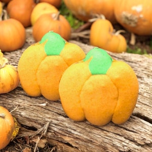 May include: Two orange pumpkin-shaped decorations with green stems sit on a weathered wooden surface, surrounded by other pumpkins. The pumpkins have a textured surface and are likely for fall or Halloween decor.