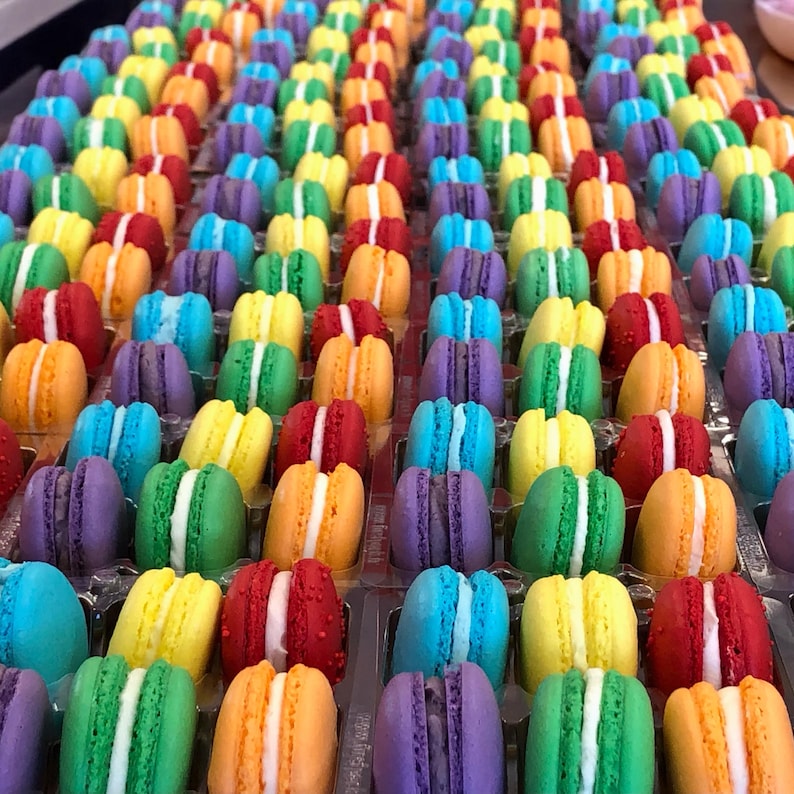 May include: A large collection of colorful macarons arranged in a grid pattern. Each macaron features two round cookies in shades of red, orange, yellow, green, blue, and purple, sandwiching a thick layer of white cream filling in the center.