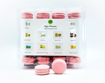 May include: A clear plastic box filled with twelve pink vegan macarons. The macarons are arranged in a grid pattern. The box has a white label with the text "Vegan Macaron" and the website "veganmacaronstore.com". The label also lists the flavors of the macarons: Birthday, Amaretto, Temero Rocher, Almond, Masala Chai, Chocofite, Cotton Candy, Banana Peanut Fudge, Vanilla Sea Salt, and Mango Coconut.