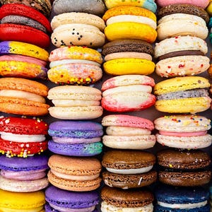 May include: A collection of colorful French macarons arranged in a grid pattern. The cookies feature various shades including red, yellow, blue, purple, brown, and beige, each with a visible cream filling sandwiched between two round, textured shells.