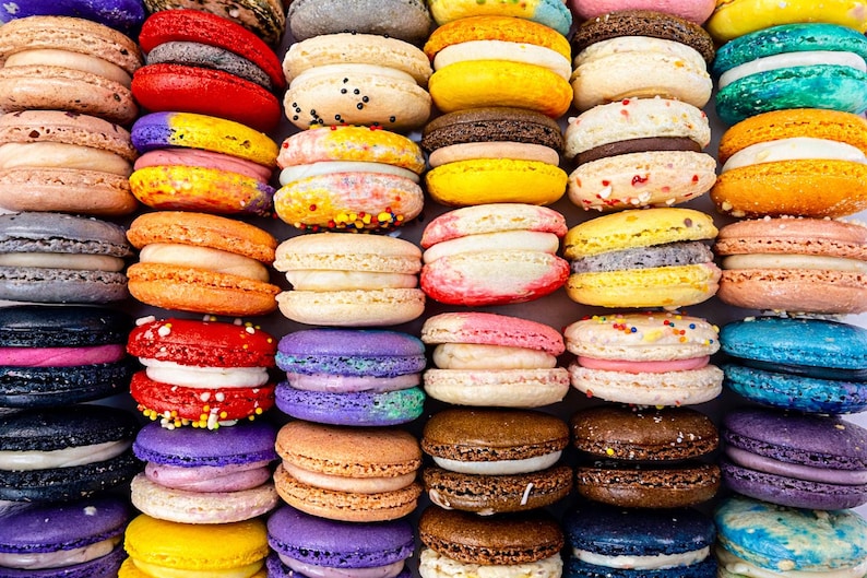 May include: A collection of colorful French macarons arranged in a grid pattern. The cookies feature various shades including red, yellow, blue, purple, brown, and beige, each with a visible cream filling sandwiched between two round, textured shells.