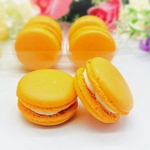 May include: A close-up of six orange macarons, two of which have white filling. The macarons are arranged in a row on a white surface.