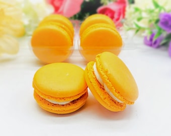 Può includere: Primo piano di sei macarons arancioni, di cui due con ripieno bianco. I macarons sono disposti in fila su una superficie bianca.