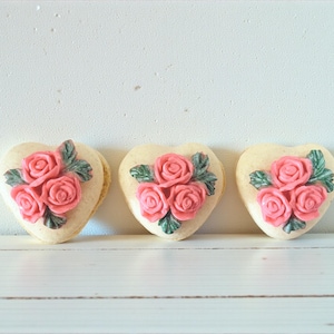May include: Three heart-shaped cookies with pink rose decorations and green leaves. The cookies are light-colored and sit on a white surface. The roses are a vibrant pink, and the leaves are a muted green.
