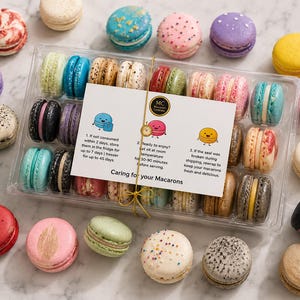 May include: A clear plastic box filled with assorted colorful macarons, surrounded by loose macarons in shades of red, blue, yellow, purple, and white. A white instruction card titled Caring for your Macarons is tied to the box with gold string.