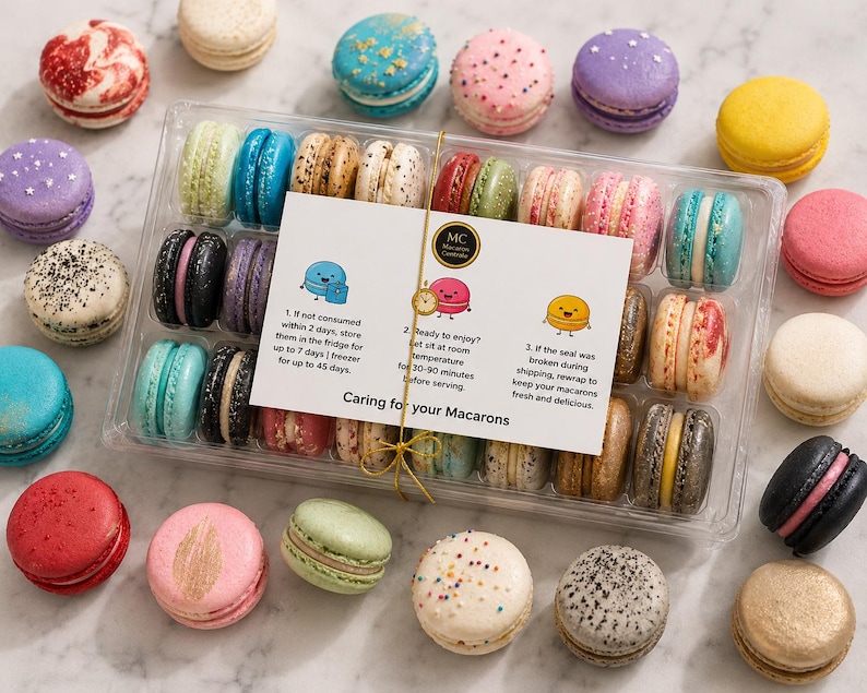 May include: A clear plastic box filled with assorted colorful macarons, surrounded by loose macarons in shades of red, blue, yellow, purple, and white. A white instruction card titled Caring for your Macarons is tied to the box with gold string.