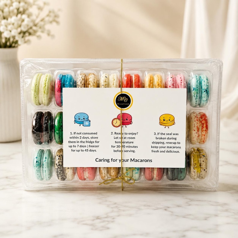 May include: A clear plastic rectangular container filled with twenty-four assorted colorful macarons, arranged in three rows. A white instructional card titled Caring for your Macarons is centered on the front, featuring three cartoon illustrations and text detailing storage and serving instructions.