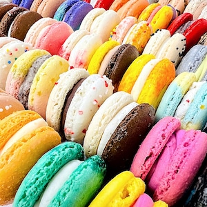 May include: A collection of colorful macarons arranged in rows, featuring various shades including pink, yellow, green, blue, brown, and white. Many of the round sandwich cookies have visible fillings, sprinkles, or textured surfaces, creating a vibrant display.
