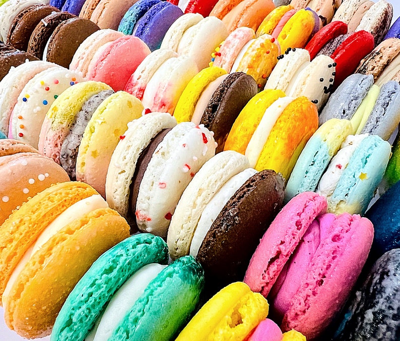 May include: A collection of colorful macarons arranged in rows, featuring various shades including pink, yellow, green, blue, brown, and white. Many of the round sandwich cookies have visible fillings, sprinkles, or textured surfaces, creating a vibrant display.