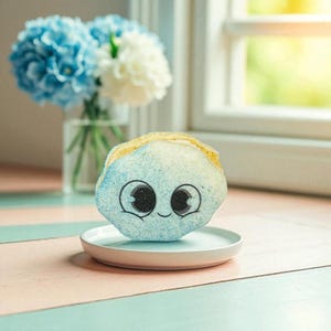 May include: A blue and yellow bath bomb shaped like a cartoon character with large eyes and a smiling mouth, resting on a small white plate. A vase of blue and white flowers is in the background.