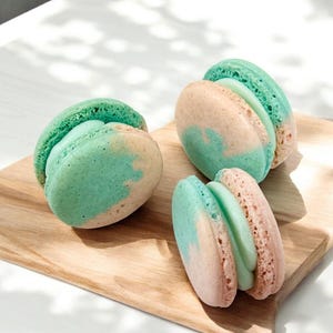 May include: Three colorful macarons on a wooden board. The macarons have a light pink and teal color scheme. The filling is a light green color. The macarons are arranged on a light brown wooden board.