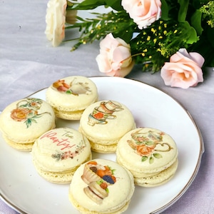 May include: Six pastel-colored macarons decorated with images of teacups and the text "Tea Party!" on a white plate.