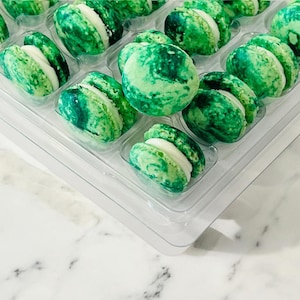 May include: A clear plastic tray filled with green macarons. The macarons have a marbled green appearance with a white filling. The macarons are arranged in rows and are a sweet treat.
