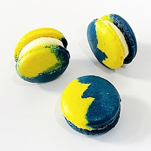 May include: Three yellow and blue macarons with white filling. The macarons are arranged in a triangular shape on a white surface.