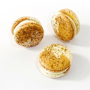 May include: Three golden brown macarons with white filling. The macarons are dusted with a light brown powder.