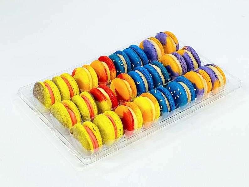 May include: A clear plastic container filled with colorful macarons. The macarons are arranged in rows, with each row featuring a different color: purple, blue, yellow, orange, and red. The macarons are all round and have a smooth, glossy finish.