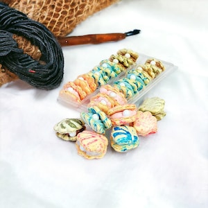 May include: A clear plastic container filled with colorful macarons shaped like seashells. The macarons are decorated with white pearl candies and have a variety of blue, pink, and yellow designs.