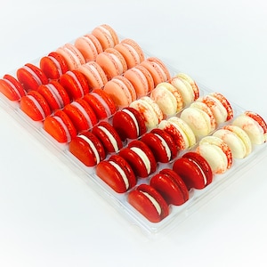 May include: A clear plastic box filled with 36 macarons. The macarons are arranged in three rows of 12. The macarons are in shades of pink, red, and white.