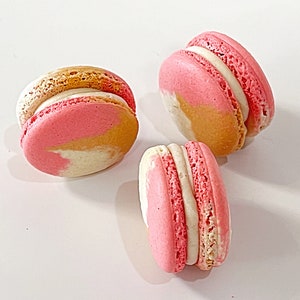May include: Three pink and gold macarons with white filling. The macarons are arranged in a triangle shape on a white surface.