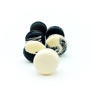 May include: An assortment of macarons in various colors and designs. Some macarons are black with white accents, while others have a marbled appearance in shades of black and cream. One macaron is a solid cream color.