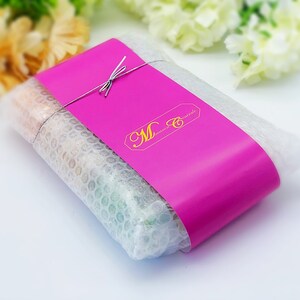 May include: A pink package wrapped in bubble wrap and tied with a string. The package has a pink label with the text "M Creations" in gold lettering.