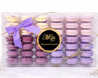 May include: A clear plastic box filled with colorful macarons. The macarons are arranged in rows and columns, with shades of purple, pink, and yellow. A purple ribbon and a gold-colored wire are tied around the box. The box has a label that says "Macaron Centrale".
