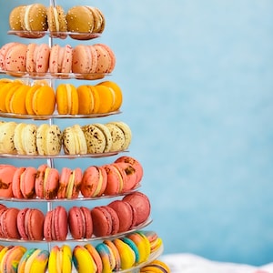 May include: A tower of colorful macarons on a clear tiered stand. The macarons are arranged in rows and are a variety of colors, including pink, yellow, and blue. The macarons are a popular French dessert.