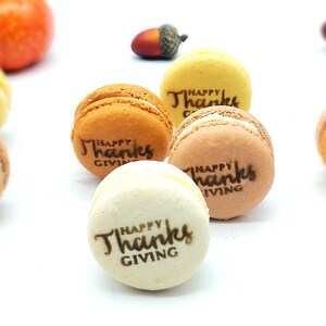 May include: An assortment of colorful macarons with the text "Happy Thanksgiving" printed on them. The macarons are in shades of orange, yellow, and brown, with a white filling. Decorative gourds and acorns are in the background.