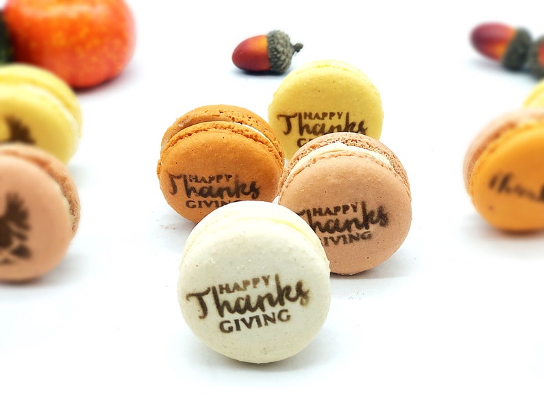 May include: An assortment of colorful macarons with the text "Happy Thanksgiving" printed on them. The macarons are in shades of orange, yellow, and brown, with a white filling. Decorative gourds and acorns are in the background.
